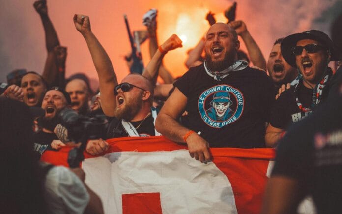 Njaveći derbiji Bakana. Fudbal. Navijači. Fudbalski navijači pjevaju dok idu na utakmicu držeći zastavu i upaljene baklje.