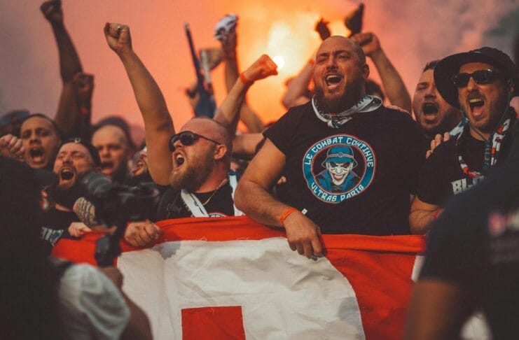 Njaveći derbiji Bakana. Fudbal. Navijači. Fudbalski navijači pjevaju dok idu na utakmicu držeći zastavu i upaljene baklje.