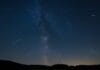 Astronomski događaji 2025, Noćno nebo sa kišom meteora tokom Perzeida iznad mirnog pejzaža, uz vidljivu Mliječnu stazu i siluete drveća.