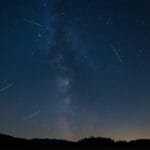 Astronomski događaji 2025, Noćno nebo sa kišom meteora tokom Perzeida iznad mirnog pejzaža, uz vidljivu Mliječnu stazu i siluete drveća.