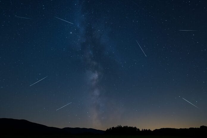 Astronomski događaji 2025, Noćno nebo sa kišom meteora tokom Perzeida iznad mirnog pejzaža, uz vidljivu Mliječnu stazu i siluete drveća.