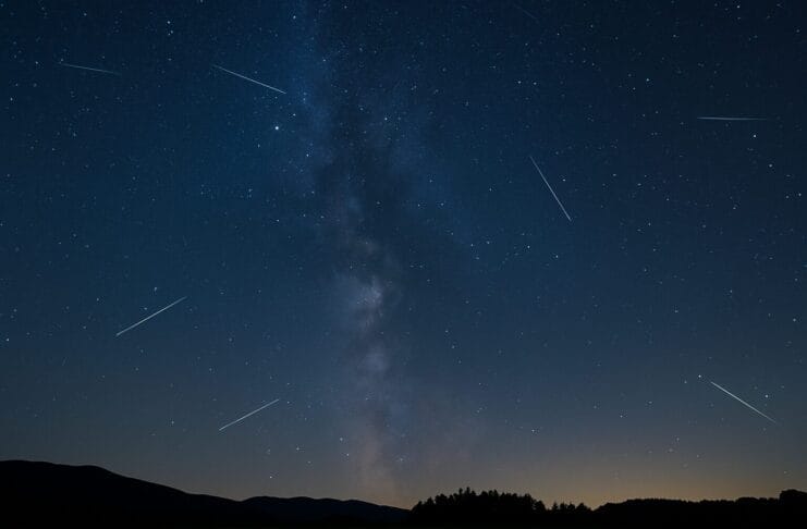 Astronomski događaji 2025, Noćno nebo sa kišom meteora tokom Perzeida iznad mirnog pejzaža, uz vidljivu Mliječnu stazu i siluete drveća.
