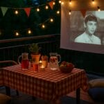 Ljetno kino na balkonu s lampicama, bokalom s voćem, jastučićima i projekcijom filma u toploj večeri