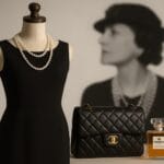 Mala crna haljina na manekenu s biserima, uz Chanel torbicu i parfem No. 5, u pozadini zamućena crno-bijela fotografija Coco Chanel.
